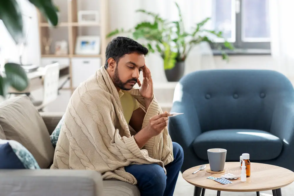 people, health and fever concept - sick indian man in blanket measuring temperature by thermometer at home